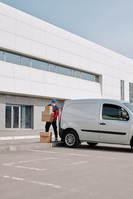 A person wearing a blue beanie and red jacket is seen carrying three cardboard boxes, one in each hand and one under the arm, as they load them into the back of a light grey van parked outside a modern white building with large windows. The scene takes place on a paved parking area with marked parking spaces, indicating an organized transport or home relocation process. The individual is positioned near the building's entrance, possibly involved in packing or furniture transport, which aligns with house removals and moving services offered by Man with Van Merton Park. The image captures the loading process during daylight with natural lighting, emphasizing the practical aspects of packing and moving logistics, including unpacking supplies, loading equipment such as trolleys or straps, and the careful handling of household items for a home relocation.