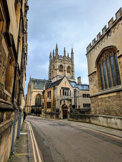 A narrow urban street scene featuring historic stone buildings with large, arched windows on either side, leading towards a prominent medieval church with a tall, detailed tower topped with spires. The street is paved with asphalt and has a double yellow line along the curb, indicating parking restrictions. The buildings exhibit Gothic architectural elements, such as pointed arches and ornate stonework, and are constructed from weathered, light-colored stone. There are no vehicles or pedestrians visible, and the sky overhead is partly cloudy with patches of blue. This setting reflects a typical location where home relocation or furniture transport might take place, with the church serving as a notable landmark in a historic area. [COMPANY_NAME] offers removals services to facilitate house moves within such environments, ensuring careful transport of furniture and belongings through city streets.