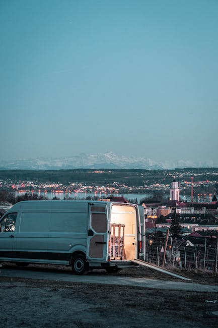 A person wearing a blue beanie and red jacket is seen carrying three cardboard boxes, one in each hand and one under the arm, as they load them into the back of a light grey van parked outside a modern white building with large windows. The scene takes place on a paved parking area with marked parking spaces, indicating an organized transport or home relocation process. The individual is positioned near the building's entrance, possibly involved in packing or furniture transport, which aligns with house removals and moving services offered by Man with Van Merton Park. The image captures the loading process during daylight with natural lighting, emphasizing the practical aspects of packing and moving logistics, including unpacking supplies, loading equipment such as trolleys or straps, and the careful handling of household items for a home relocation.