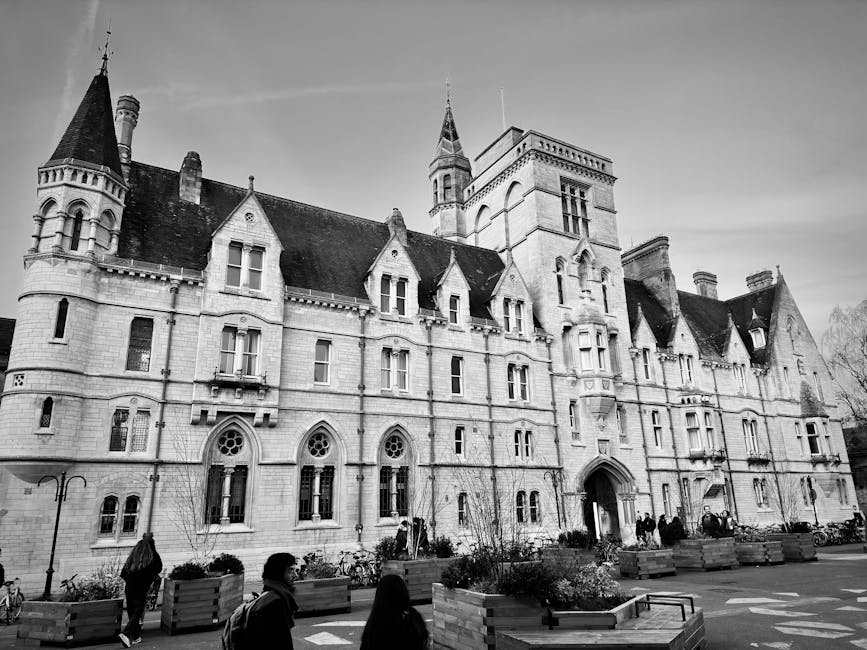 Black and white photograph of a large, historic castle-style building with multiple turrets, pointed spires, and tall, narrow windows, situated in an urban area with a paved street and pedestrian walkway in the foreground. Several people are walking or standing near the entrance, which features an arched doorway. The sidewalk is lined with rectangular wooden planters containing small plants and trees. On the street, bicycles are parked, and a few individuals are visible near the building. The image captures the exterior of the property during daytime, with natural lighting and clear weather conditions, illustrating the type of architecture that might be associated with residential or institutional properties requiring professional removals and home relocation services, as provided by Man with Van Merton Park, with an emphasis on loading and transport considerations for furniture and packing materials.