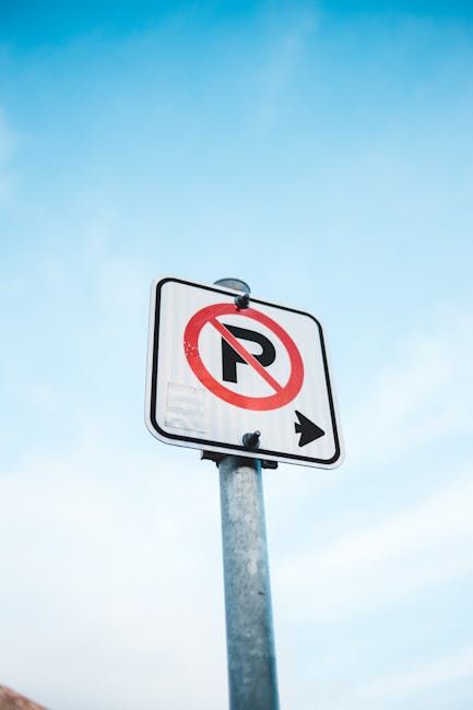 A rectangular traffic sign mounted on a metal pole, displaying a red circle with a diagonal line crossing out the letter 'P' to indicate no parking, with a black arrow pointing to the right below. The sign is positioned outside—likely on a pavement—against a backdrop of a clear blue sky. The image captures the sign at a slight upward angle, emphasizing the prohibition message. This visual relates to parking restrictions that may impact home relocation or moving logistics, important considerations for services like those offered by Man with Van Merton Park during furniture transport and removal processes.