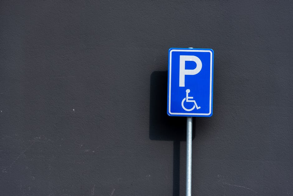 A blue parking sign with a white 'P' and a wheelchair symbol indicating accessible parking, mounted on a silver metal post against a plain dark grey wall. The sign is outdoors, and the lighting creates a shadow of the post on the wall. The image captures the sign in a close-up view, with no other objects visible around it. This type of accessible parking permit sign is relevant in the context of house removals or relocation services, where special parking arrangements may be necessary for vehicle access during moving operations, with the sign positioned near the property entrance or on the pavement.
