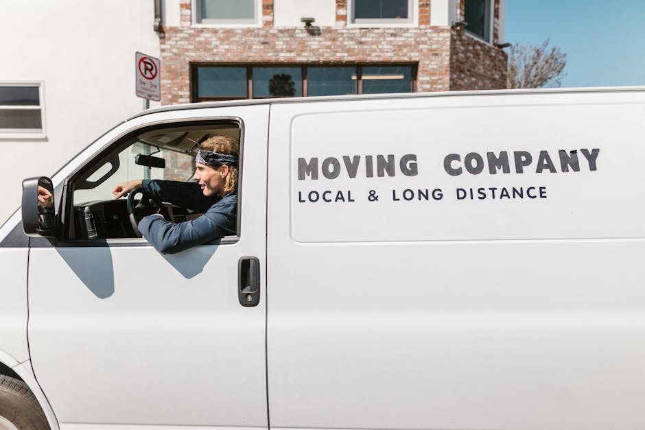 A white moving van parked outside a residential building with a brick facade, displaying the company name 'Man with Van Merton Park' and the service category 'removals' on its side. The side door of t