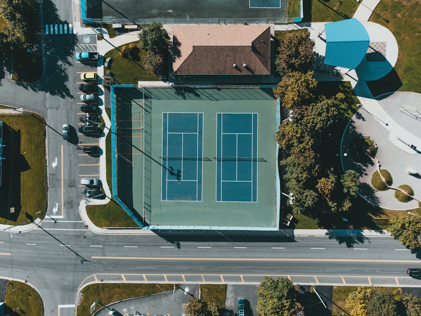 Aerial view of a sports court surrounded by trees, residential buildings, and parking areas. The court features two standard tennis or basketball courts with marked lines, enclosed by a high fence. Adjacent to the court, there is a small blue pavilion with a curved roof, situated on a paved circular area. Nearby, a group of parked cars is visible along the street, with some situated on a designated parking lane. The environment is well-lit with natural daylight, highlighting the tidy, organized setting. This setting exemplifies a typical outdoor recreational space near residential homes, such as those managed or accessed during house removals or home relocation services provided by Man with Van Merton Park.