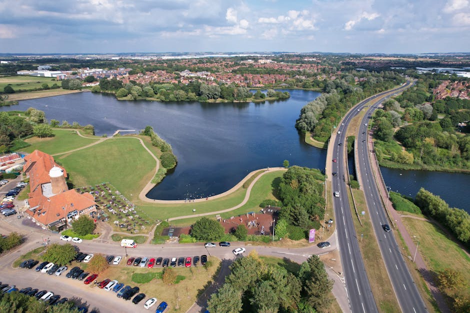 An aerial view showing a large body of water with surrounding green parkland and pathways, adjacent to a residential area with houses and trees. A major dual carriageway with several vehicles passes alongside the park, leading towards the horizon. In the foreground, a building with a red-tiled roof and a parking lot filled with cars is visible. The scene depicts a peaceful suburban environment with natural and built features, illustrating a typical setting for house removals or property relocation services provided by Man with Van Merton Park. This image captures the logistics of home relocation, including transport routes and accessible green spaces that are often involved in furniture transport and packing and moving procedures.