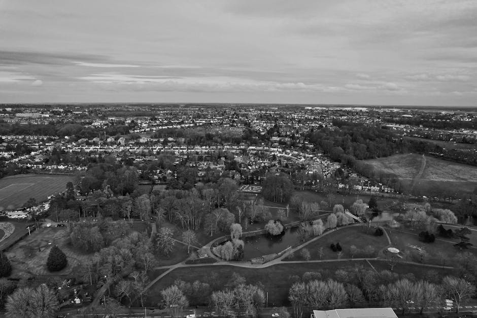 A black and white aerial photograph showing a residential area with a large park in the foreground, featuring walking paths, trees, and a small pond. Surrounding the park are rows of houses and streets stretching into the distance, with some open fields and patches of greenery visible beyond the built-up area. The image captures a wide view of Merton Park during daytime with natural lighting conditions. This scene illustrates a typical urban environment where [COMPANY_NAME], such as Man with Van Merton Park, facilitates home relocations by enabling the safe loading and transport of furniture, boxes, and packing materials via vans positioned near the property, reflecting the logistics involved in a same-day moving service.