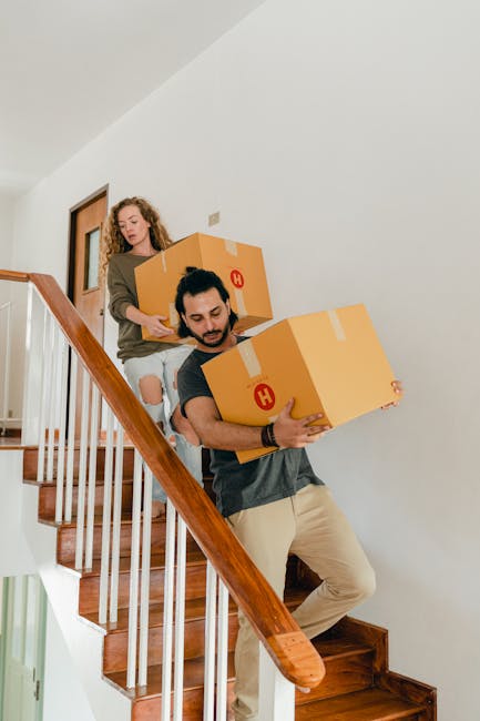 Two professional movers from Man with Van Merton Park are carrying cardboard boxes down a wooden staircase during a house removal. The man in the foreground is holding a large cardboard box labeled with a red circle containing a white 'H', while the woman behind him is guiding another box following closely. The staircase has wooden steps and a wooden handrail, with white-painted walls surrounding it. The scene is well-lit with natural light, and the movers are wearing casual clothing suitable for packing and moving activities. This image depicts the careful handling and transportation of boxes as part of home relocation services, focusing on the loading process within a residential property, aligned with the house removals and moving services offered by [COMPANY_NAME].