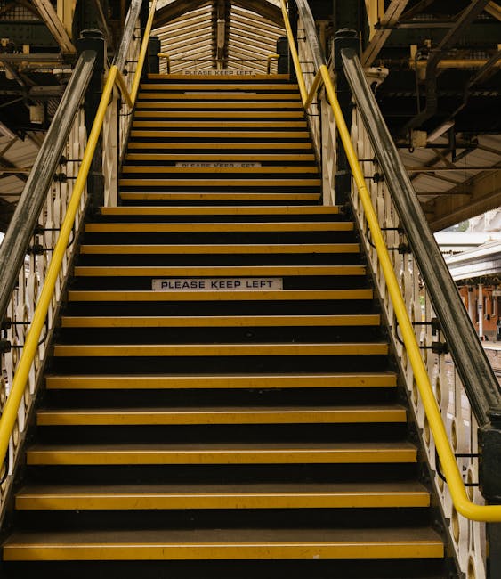 A staircase inside a building with metal railings and yellow safety bars on both sides, leading up to a mezzanine level. The steps are made of wood with black anti-slip strips on the edges. A white sign with black text reading 'PLEASE KEEP LEFT' is affixed to one of the middle steps. The surrounding environment includes industrial structural elements, such as metal beams and pipes, and a partially visible loading area beneath the stairs, indicating an industrial or warehouse setting. The lighting is utilitarian, with overhead fixtures illuminating the stairway, which is part of a facility possibly used for logistics or storage, supporting the context of home relocation and furniture transport services offered by Man with Van Merton Park.